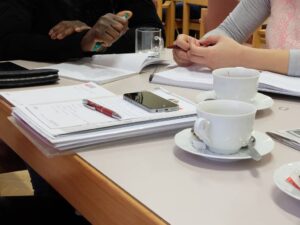 Zwei Personen sitzen an einem Tisch mit Arbeitsunterlagen und Kaffeetassen.